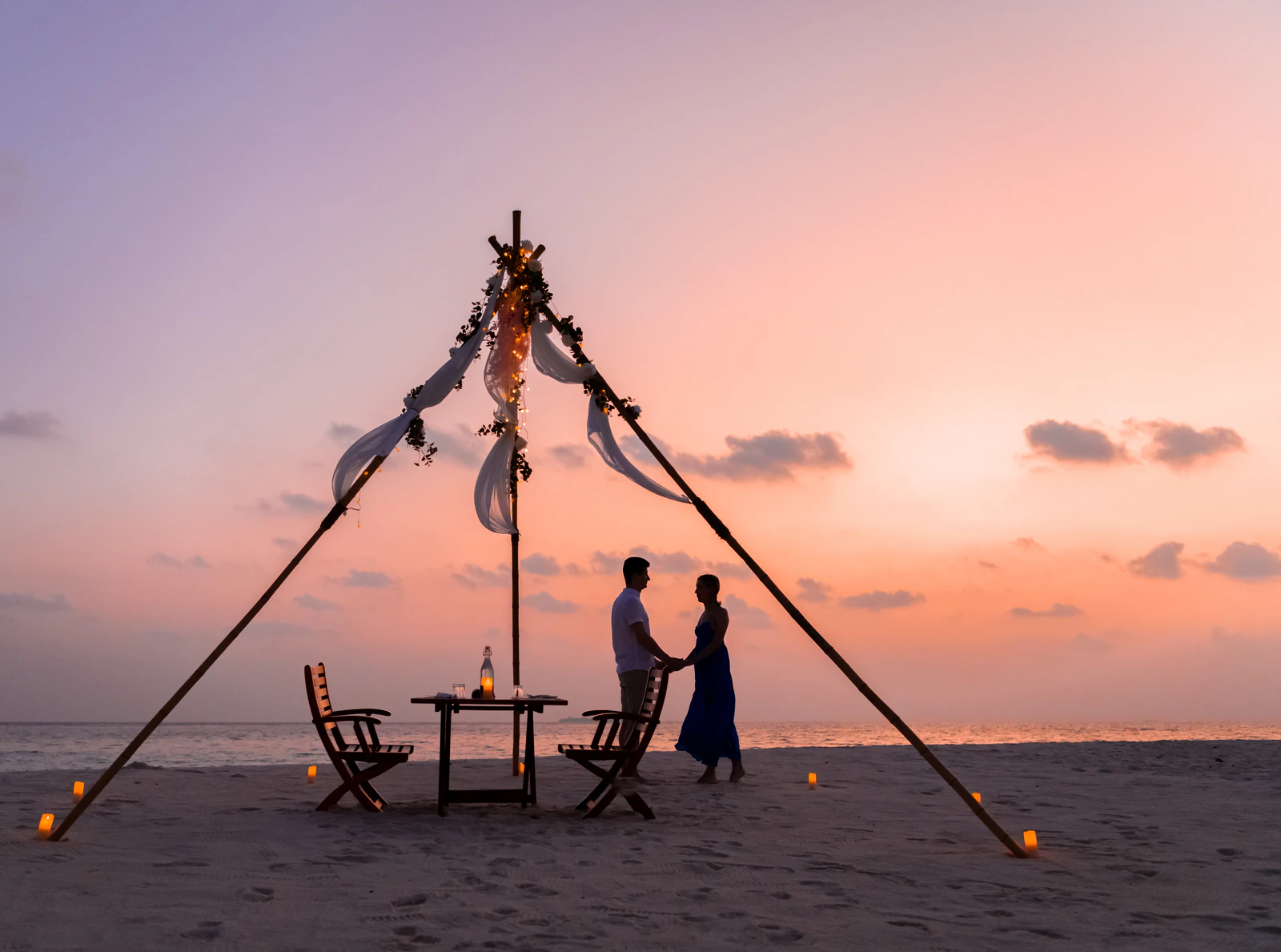 Private beach dinner setup with a couple beneath a decorated canopy at sunset